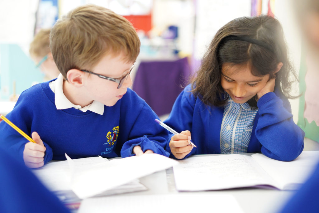 Two young Dartford Primary Academy students are shown sat at their desks and smiling whilst holding pens in their hands and looking at their work.