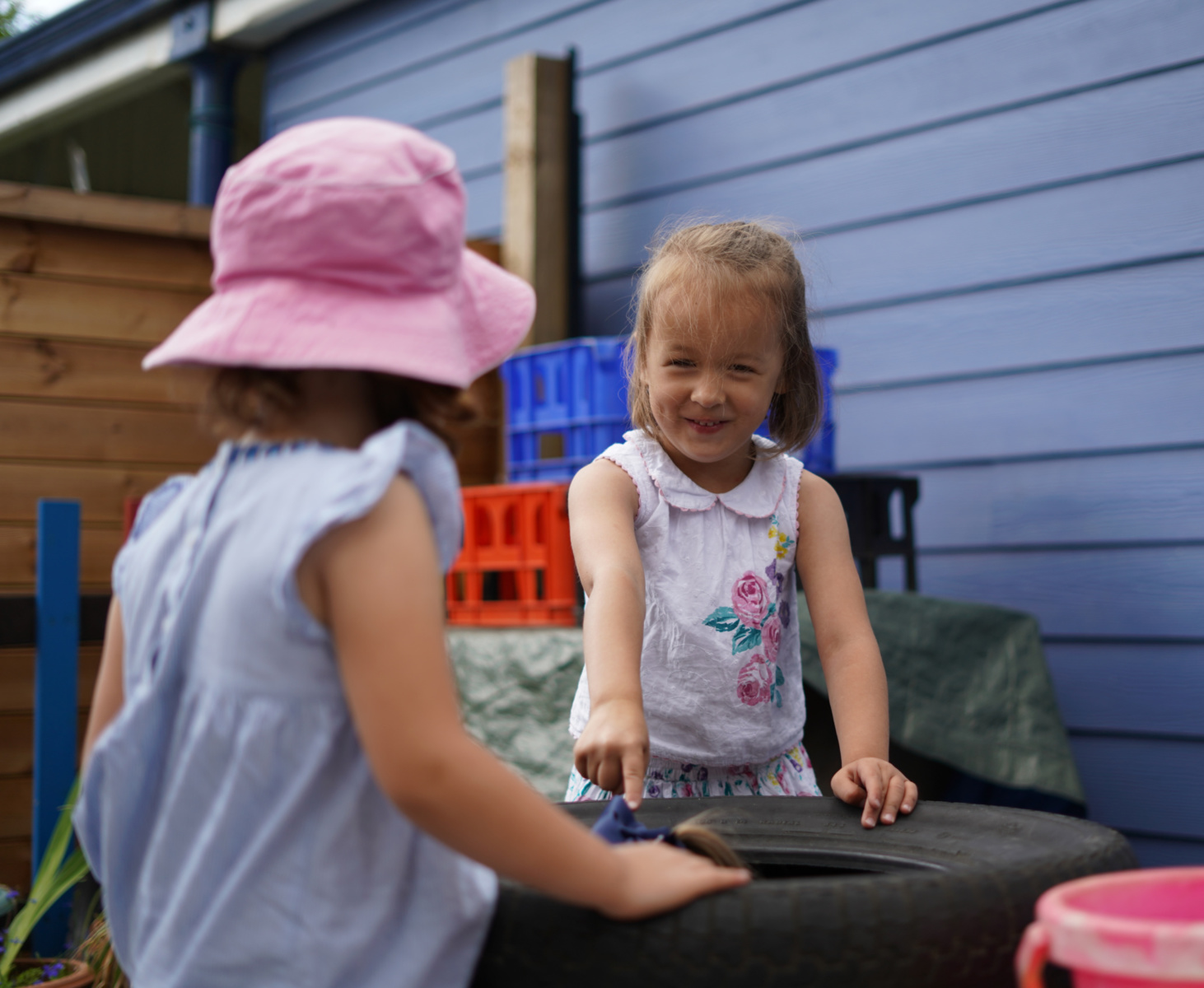 Two young Nursery-aged girls are pictured outdoors on the academy grounds, smiling at one another. One of the girls is seen pointing to something inside a car tyre.