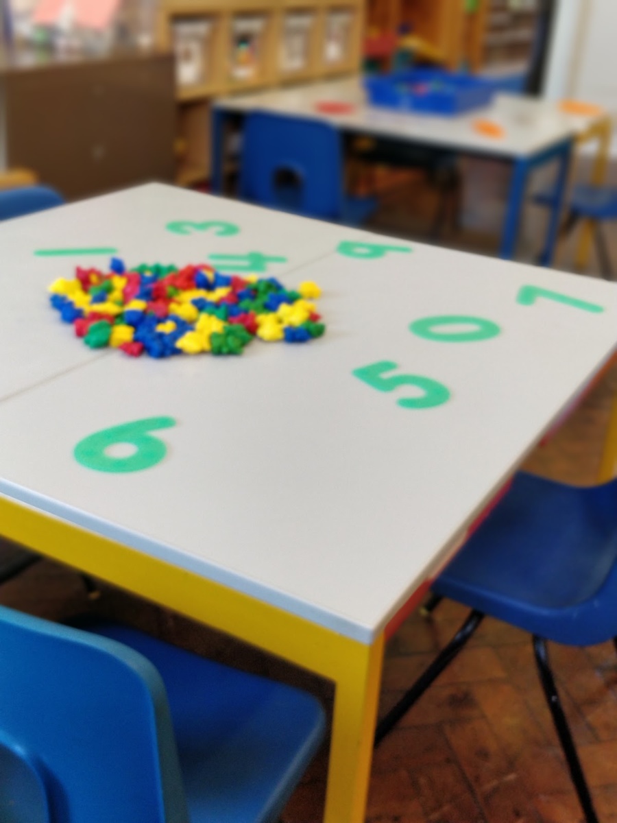 A photo of a table in the middle of a classroom. On top of it are numbers and multi-coloured counters laid out.