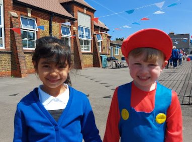 Two children, a boy and a girl, are seen on the academy playground, smiling for the camera together. The boy is dressed up as Mario from the Nintendo video game.