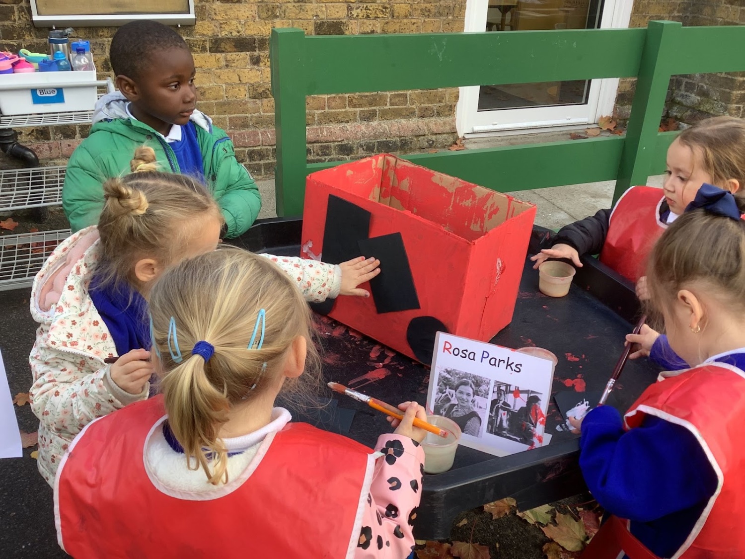 A small group of pupils are seen outdoors on the academy grounds, decorating a box using paints and coloured paper.