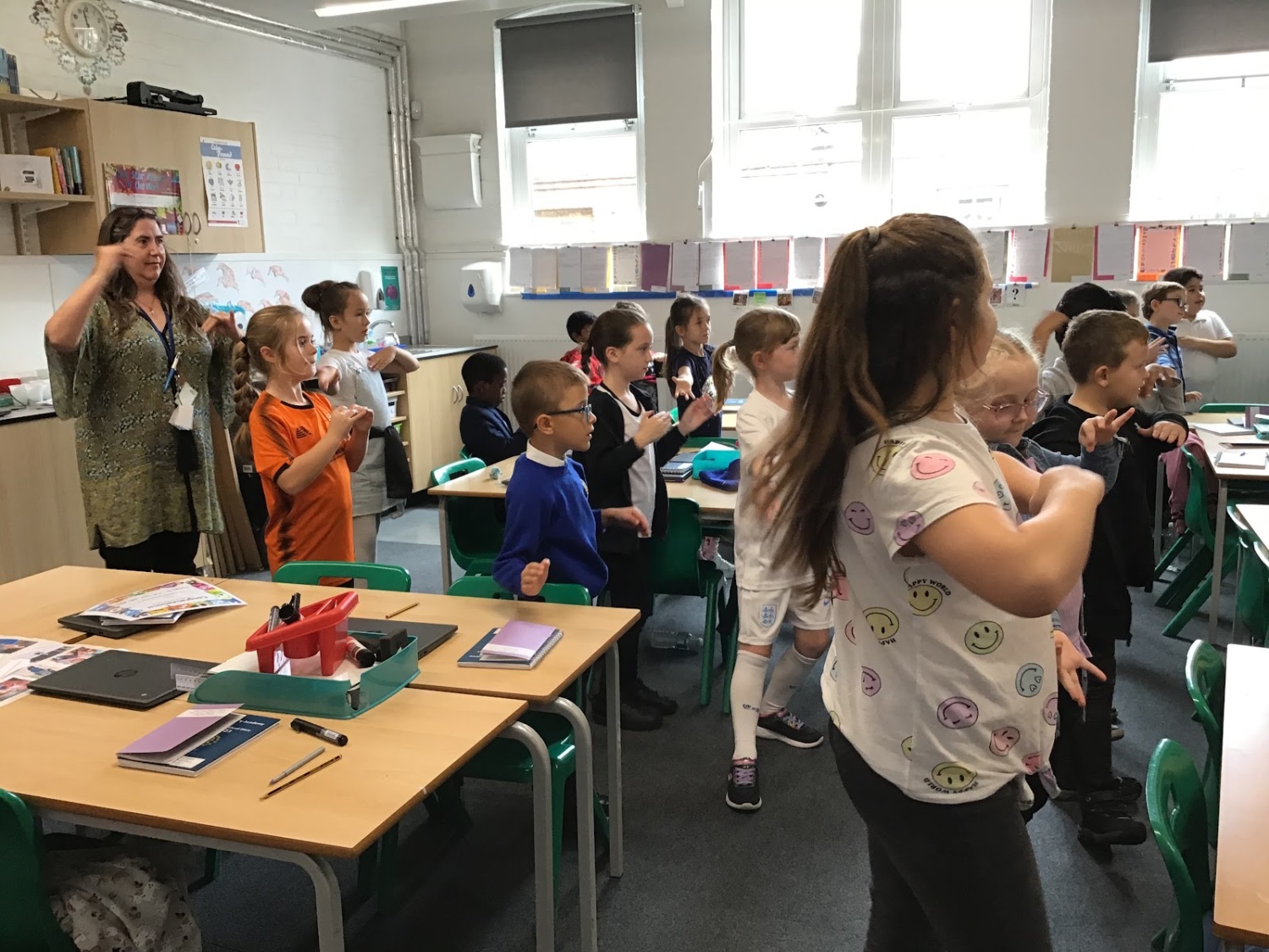 A class of young pupils are pictured stood behind their chairs in their classroom, participating in a dance activity, whilst under the supervision of a member of staff.