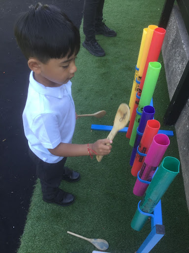 A young boy from Nursery can be seen experimenting with different coloured tubes and beating against them with a wooden spoon to hear the sounds that they make.