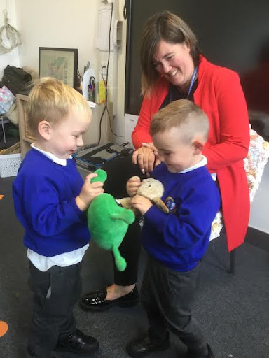 Two young boys are shown playing with soft toys together in a classroom, under the supervision of a member of staff.