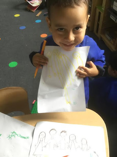 A young boy in Nursery is seen smiling at the camera, whilst showing a drawing he has created in class.