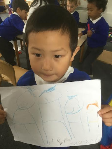 A young boy in Nursery is seen smiling at the camera, whilst showing a drawing he has created in class.