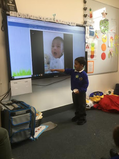 A young boy is seen standing at the front of the Nursery class and pointing at the Interactive Board, presenting to his peers.