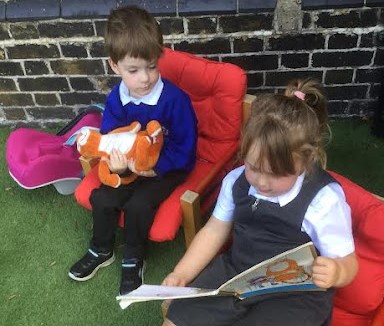 Two young Nursery pupils, a boy and a girl, are seen sat together outdoors on the school grounds. The girl is reading a storybook called 'The Tiger Who Came To Tea'. The boy is holding a Tiger soft toy in his hands.