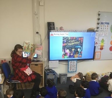 A class teacher is seen sat at the front of the classroom, reading a storybook to her class who are sat on the floor in front of her.