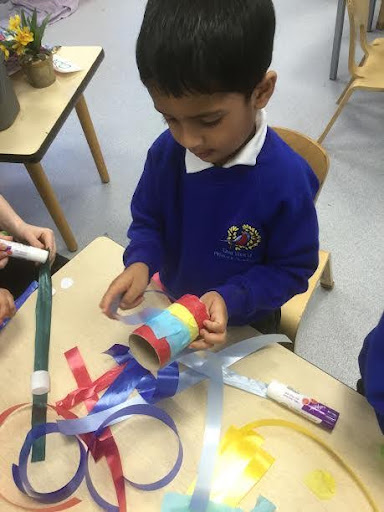 A young boy is seen sat at his desk in a classroom, decorating a toilet roll tube with some coloured paper and craft materials.