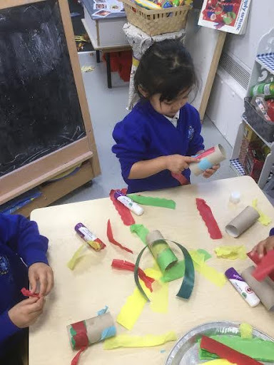 A young girl is seen sat at her desk in a classroom, decorating a toilet roll tube with some coloured paper and craft materials.