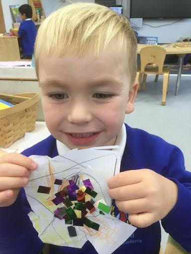 A young boy from Nursery can be seen smiling for the camera, whilst showing a piece of artwork he has created, using glue and coloured paper.