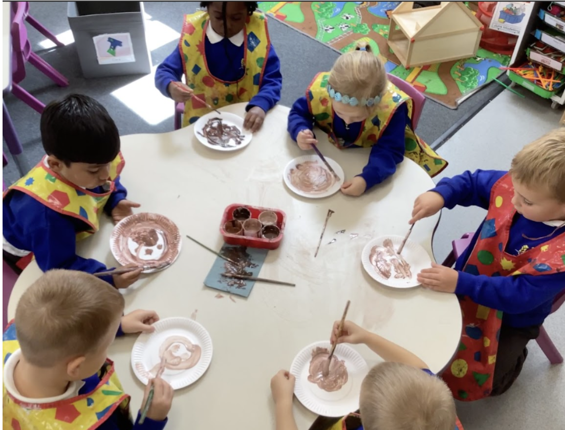 Six pupils are pictured sat around a table, wearing their art aprons, painting paper plates with brown coloured paint.