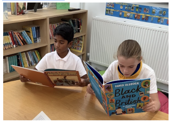 Two young pupils, a girl and a boy, are pictured sat at a desk, wearing their academy uniform and reading books on the topic of Black History.
