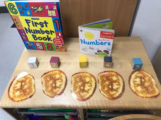 Five cooked Chapattis are seen laid out on a wooden desk in a classroom with numbered blocks above them, counting them from one to five. Two books on number counting are also shown on the desk.