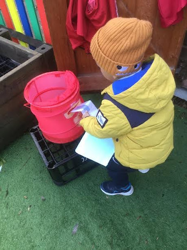 A young boy from Nursery is seen outdoors, wearing his winter coat, posting a Christmas card he has made through a postbox.
