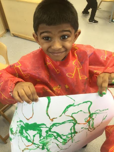 A young boy from Nursery is pictured smiling for the camera, whilst wearing an art apron and showing a painting he has made.