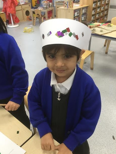 A young girl from Nursery is seen smiling for the camera and wearing a hat she has made from card.
