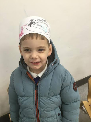 A young boy from Nursery is seen smiling for the camera and wearing a hat he has made from card.
