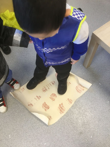 A young boy in Nursery can be seen creating footprints with his shoes, using paint on a sheet of paper on the floor.