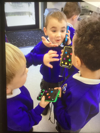 A small group of young boys are seen playing with toy mobile phones together in their classroom and smiling at one another.