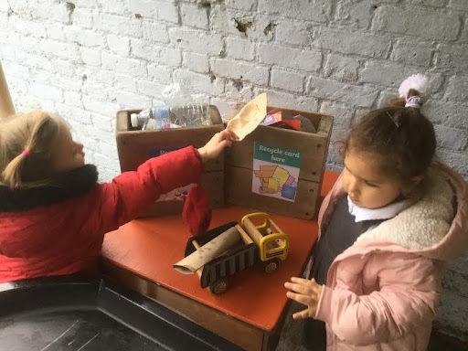 Two young girls from Nursery are pictured outdoors in their winter coats, putting different junk items into recycling bins.