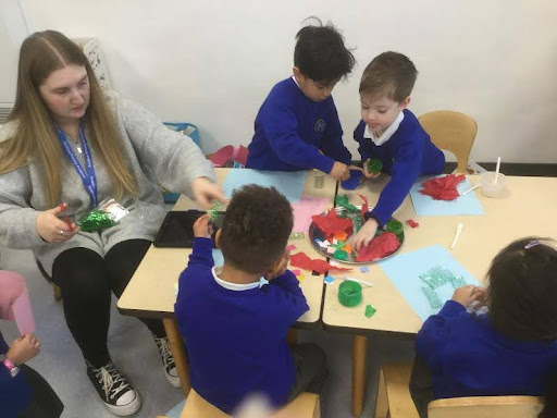Four Nursery pupils are seen creating their own collages on paper with craft materials, under the supervision of a member of staff who is sat with them.