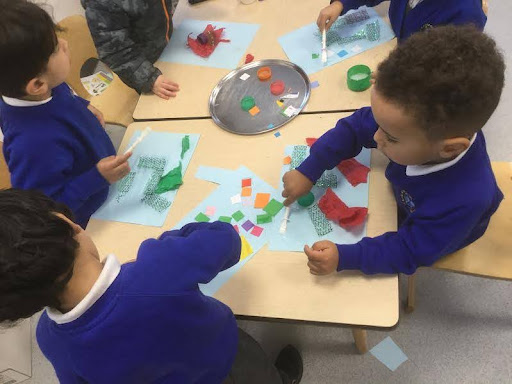 Five Nursery pupils are seen sat together at a table, creating their own collages on paper with craft materials.