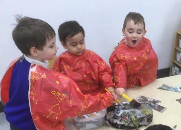 Three young boys from Nursery are shown wearing art aprons and using PVA glue to stick together newspaper in order to create a model.
