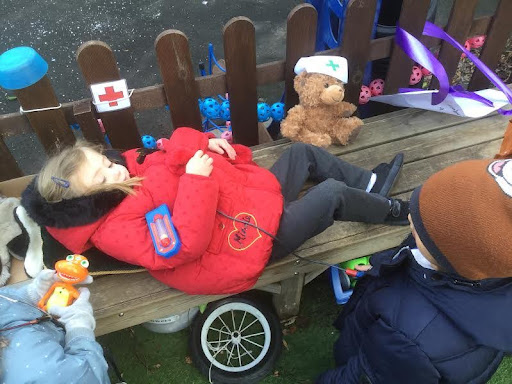 A young girl from Nursery is pictured laying down on a wooden bench outdoors, pretending to be a patient seeing the doctor.