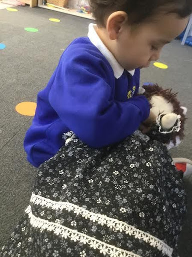 A young boy in Nursery is pictured sitting on the floor in a classroom, holding a dolly in his hands and cuddling it.
