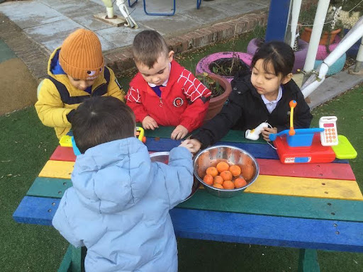 Three young Nursery pupils are pictured standing behind a bench, pretending to sell items at a shop. They are seen interacting with another pupil pretending to be a customer on the other side of the bench.