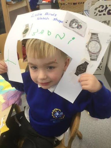 A young boy from Nursery is pictured holding up an advert for wristwatches he has designed on paper for the camera.
