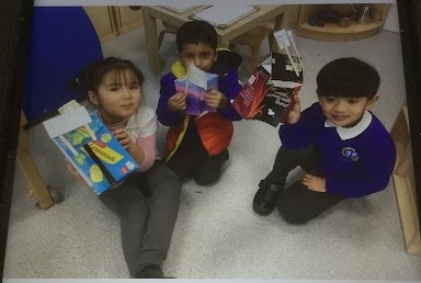 Three young pupils from Nursery are seen sat on the floor, smiling for the camera and holding up different kinds of food packaging they have found.