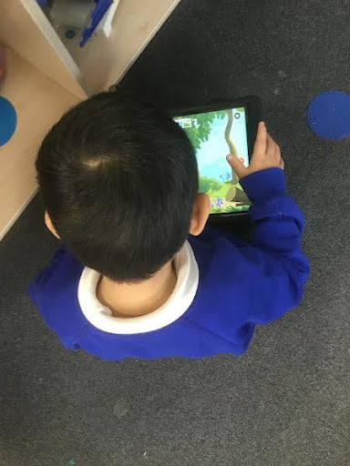 A young boy from Nursery is seen from behind, playing a game on a tablet computer, whilst sitting on the floor in his classroom.