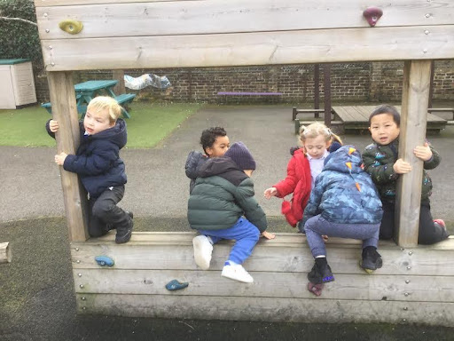 A group of six Nursery pupils can be seen climbing on a wooden apparatus, outdoors on the academy grounds.