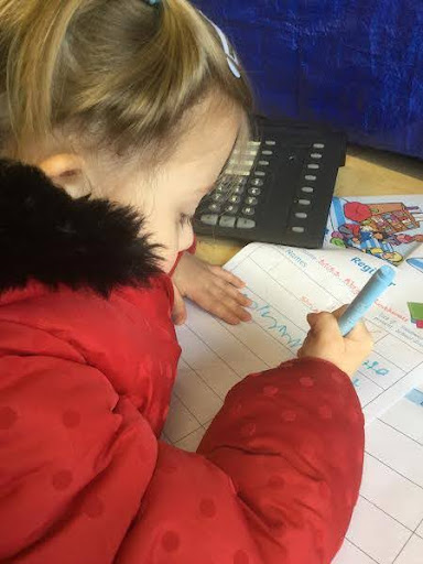 A young girl from Nursery is shown sat at her desk, completing a worksheet and wearing her winter coat.