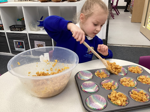 A young girl from Reception is pictured spooning cake mixture from a large plastic bowl into individual cake cases on a tray.