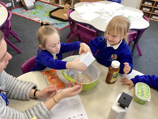 Two female pupils from Reception are seen mixing ingredients together to make a cake, whilst under the supervision of a staff member.