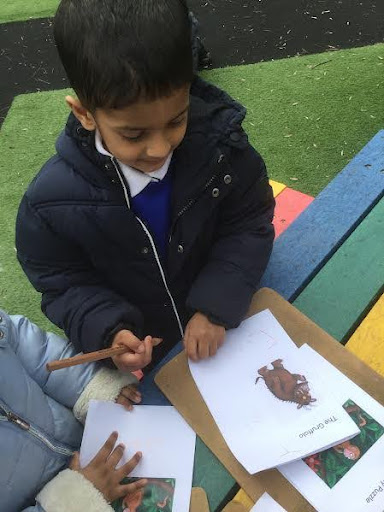 A young boy from Nursery is pictured wearing his winter coat outdoors on the academy grounds, whilst drawing pictures of characters from 'The Gruffalo' on paper.