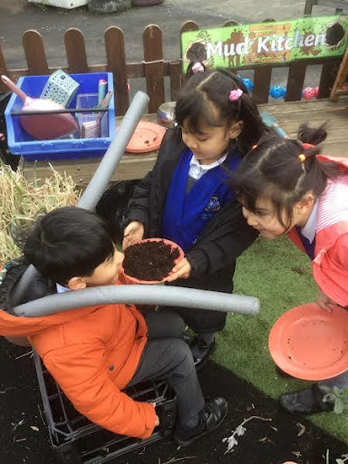A group of three Nursery pupils can be seen sat together outdoors, playing with some soil they have put into a plant pot.