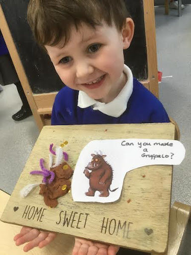 A young boy from Nursery can be seen smiling for the camera, as he shows a picture of the 'Gruffalo' that he has made using craft materials on a wooden board.