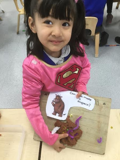 A young girl from Nursery can be seen smiling for the camera, as she shows a picture of the 'Gruffalo' that she has made using craft materials on a wooden board.