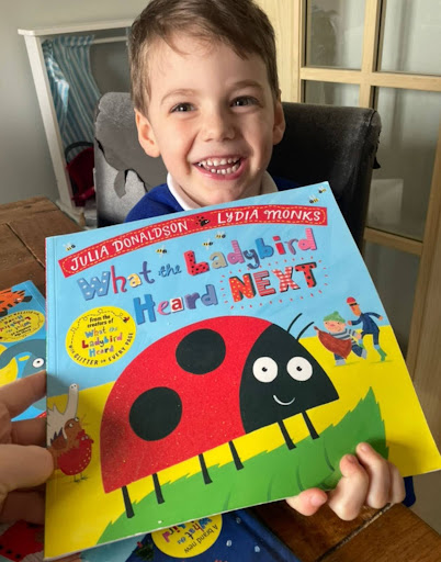 A young boy in Nursery is pictured smiling brightly for the camera, whilst holding up the storybook 'What The Ladybird Heard' by Julia Donaldson.