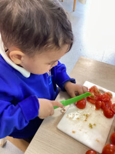 A young boy from Nursery is pictured chopping up some Tomatoes on a chopping board, using a plastic knife.