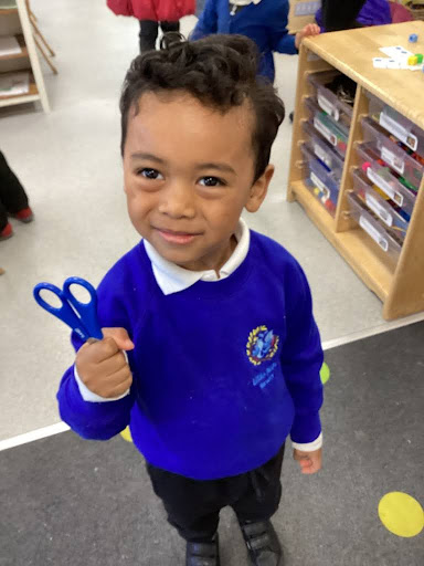 A young boy from Nursery is pictured smiling for the camera, whilst wearing his uniform and holding a pair of scissors the correct way up when walking with them.