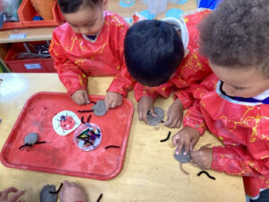 Three young pupils from Nursery are pictured creating their own Ladybird models using clay and pipe cleaners. Each of them are wearing red art aprons.