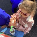 A young girl from Nursery can be seen watering some soil in a paper cup, using a watering can.