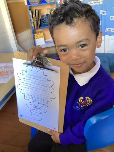 A young boy from Nursery is pictured holding up a clipboard with information he has found out about Sunflowers, whilst smiling for the camera.