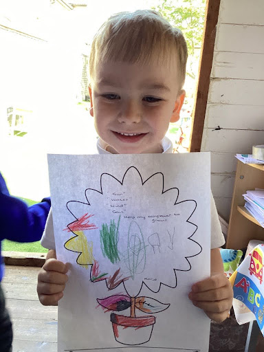 A young boy from Nursery is pictured holding up a sheet of paper with information he has found out about Sunflowers, whilst smiling for the camera.